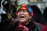 An elderly person wearing a colorful, knitted headband and traditional clothing appears to be shouting or singing. The background features people in police riot gear, adding a tension-filled atmosphere to the scene.