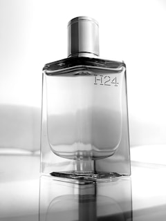 A sleek, transparent perfume bottle with the label 'H24' elegantly embossed on the front. Positioned against a minimalist, reflective surface, the bottle exhibits a modern and sophisticated design with a metallic silver cap.