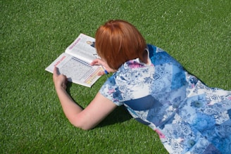 A person with reddish hair is lying on lush green grass, engaged in solving a puzzle in a magazine. They are wearing a floral-patterned dress and appear to be focusing intently on the task.