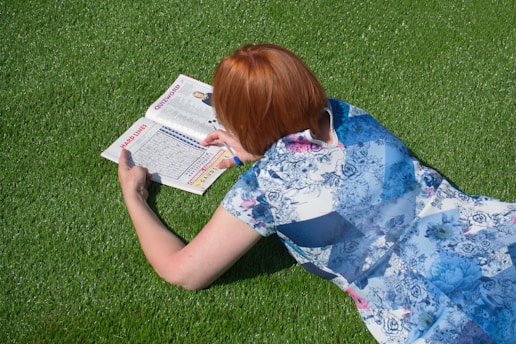 A person with reddish hair is lying on lush green grass, engaged in solving a puzzle in a magazine. They are wearing a floral-patterned dress and appear to be focusing intently on the task.