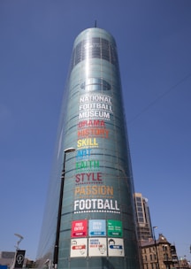 A tall cylindrical glass building displaying colorful large-font words like Drama, History, Skill, Art, Faith, Style, Passion, and Football. Free entry and open daily signs are visible at the bottom. Streetlights and a few other buildings are in the vicinity under a clear blue sky.