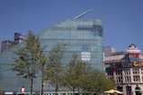 A large modern glass building with a unique angular design is prominently featured. The facade displays a sign for the National Football Museum. In the foreground, several leafy trees can be seen. To the right, various commercial signs such as The Printworks and Vue IMAX are visible, adding to the urban setting.