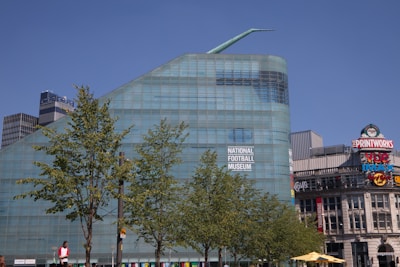 A large modern glass building with a unique angular design is prominently featured. The facade displays a sign for the National Football Museum. In the foreground, several leafy trees can be seen. To the right, various commercial signs such as The Printworks and Vue IMAX are visible, adding to the urban setting.