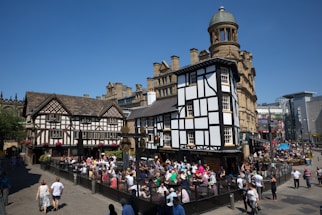 A busy outdoor scene features a large group of people sitting and socializing at tables in front of a historic half-timbered building. The buildings have a traditional architectural style with intricate woodwork and tiled roofs. People are enjoying a bright sunny day, with clear blue skies overhead.