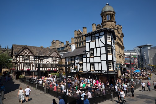 A busy outdoor scene features a large group of people sitting and socializing at tables in front of a historic half-timbered building. The buildings have a traditional architectural style with intricate woodwork and tiled roofs. People are enjoying a bright sunny day, with clear blue skies overhead.
