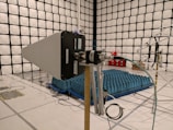 A laboratory setting featuring a large, square room with padded, soundproof walls arranged in a grid pattern. In the foreground, a large directional antenna or device with wires is positioned on a wooden stand. In the center of the room, blue foam pyramids are arranged on a platform, likely used for acoustic testing, with some red cubes and additional equipment in the background.
