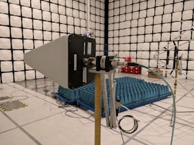 A laboratory setting featuring a large, square room with padded, soundproof walls arranged in a grid pattern. In the foreground, a large directional antenna or device with wires is positioned on a wooden stand. In the center of the room, blue foam pyramids are arranged on a platform, likely used for acoustic testing, with some red cubes and additional equipment in the background.