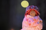 A small handcrafted doll wearing a colorful knit hat and a dress decorated with cupcake patterns. The doll's face is simple, with minimalistic features, and it is set against a dark, blurred background with a soft circular light.