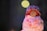 A small handcrafted doll wearing a colorful knit hat and a dress decorated with cupcake patterns. The doll's face is simple, with minimalistic features, and it is set against a dark, blurred background with a soft circular light.