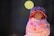 A small handcrafted doll wearing a colorful knit hat and a dress decorated with cupcake patterns. The doll's face is simple, with minimalistic features, and it is set against a dark, blurred background with a soft circular light.