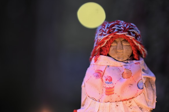 A small handcrafted doll wearing a colorful knit hat and a dress decorated with cupcake patterns. The doll's face is simple, with minimalistic features, and it is set against a dark, blurred background with a soft circular light.