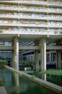 A concrete building with multiple levels of balconies adorned with plants rises above a serene water feature. Tall pillars support the structure, and the reflective water below is flanked by greenery. The architectural design features straight lines and symmetry, creating a harmonious blend with nature.