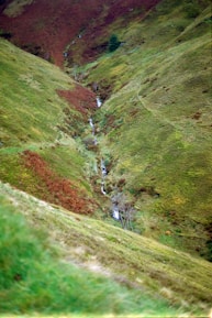 A serene landscape featuring a small waterfall cascading through a narrow, lush valley. The hills are covered in green and brown vegetation, with patches of rust-colored foliage indicating seasonal change. A lone tree stands near the waterfall, adding to the tranquil setting.