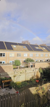A residential area with a row of brick houses featuring solar panels on the roofs. In the foreground, a fenced backyard garden is visible, containing plants, a bird bath, and a few potted plants. A rainbow is faintly visible in the sky above the houses.