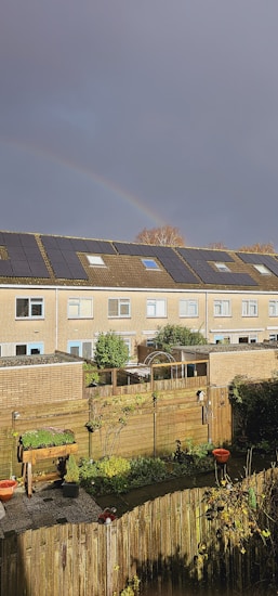 A residential area with a row of two-story houses featuring solar panels on the roofs. A rainbow arches across the overcast sky, adding a soft touch of color above the buildings. The foreground shows a tidy backyard with a wooden fence, planters, and green foliage. The atmosphere is calm and serene, typical of a suburban neighborhood.