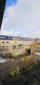 A row of modern townhouses with solar panels on their roofs, set under a partly cloudy sky. The foreground features a fenced backyard with potted plants, a small table, and patches of greenery. The scene conveys a suburban residential area with well-maintained gardens and a focus on sustainability through the use of solar energy.