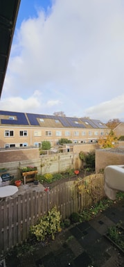 A row of modern townhouses with solar panels on their roofs, set under a partly cloudy sky. The foreground features a fenced backyard with potted plants, a small table, and patches of greenery. The scene conveys a suburban residential area with well-maintained gardens and a focus on sustainability through the use of solar energy.