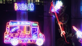 Brightly colored neon lights display a sign reading 'Bazaar' above an ice cream truck outline with various illuminated texts and illustrations. Adjacent is a neon figure holding a glowing staff, surrounded by vibrant neon patterns.
