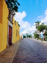 A serene cobblestone street lined with colorful colonial-style buildings. On the left, a vibrant yellow wall with a large brown wooden door is noticeable. Palm trees and trimmed bushes add greenery to the scene. The sky is a bright blue with fluffy white clouds. Flags hang above the street, and a few bicycles and park benches are visible along the sidewalks.