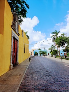 A serene cobblestone street lined with colorful colonial-style buildings. On the left, a vibrant yellow wall with a large brown wooden door is noticeable. Palm trees and trimmed bushes add greenery to the scene. The sky is a bright blue with fluffy white clouds. Flags hang above the street, and a few bicycles and park benches are visible along the sidewalks.