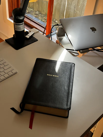 A cozy podcast recording setup with a microphone, headphones, and a Bible open on a wooden table bathed in soft morning light.
