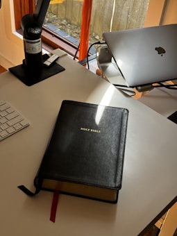A desk area featuring a closed laptop, a microphone on a stand, and a large book titled 'Holy Bible'. There is a keyboard partially visible, and sunlight is streaming in through a window, casting shadows across the desk.