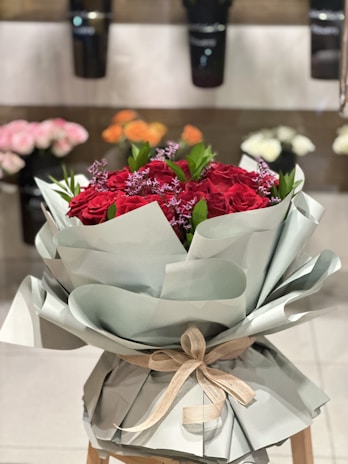 A bouquet of red roses wrapped in light green paper with a beige ribbon sits on a wooden stool. In the background, there are blurred flowers of various colors displayed in vases, suggesting a floral shop setting.