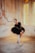 A ballet dancer is posed in a graceful position within an elegantly aged room with textured walls and soft lighting. She is dressed in a black lace ballet costume with a classic tutu, and wears pointe shoes. The ambiance is warm and vintage, with an arched window allowing a gentle glow into the room, highlighting the intricate architectural details of the walls.