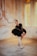 A ballet dancer is posed in a graceful position within an elegantly aged room with textured walls and soft lighting. She is dressed in a black lace ballet costume with a classic tutu, and wears pointe shoes. The ambiance is warm and vintage, with an arched window allowing a gentle glow into the room, highlighting the intricate architectural details of the walls.