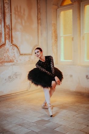 A ballet dancer is posed in a graceful position within an elegantly aged room with textured walls and soft lighting. She is dressed in a black lace ballet costume with a classic tutu, and wears pointe shoes. The ambiance is warm and vintage, with an arched window allowing a gentle glow into the room, highlighting the intricate architectural details of the walls.