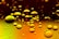A collection of various-sized oil droplets is floating on the surface of a liquid. The droplets have a reflective quality, capturing the warm hues of yellow and orange from the background.
