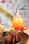 A plate of glazed ribs garnished with chopped green onions and white flowers is paired with a side of fries in a metal basket. In the background, a vibrant gradient drink in a mason jar with a straw features layers of orange, yellow, and red, styled with blueberries and a garnish.
