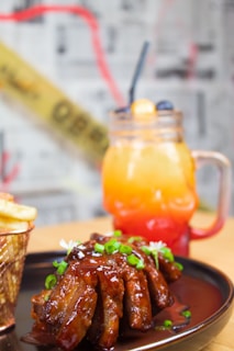 A plate of glazed ribs garnished with chopped green onions and white flowers is paired with a side of fries in a metal basket. In the background, a vibrant gradient drink in a mason jar with a straw features layers of orange, yellow, and red, styled with blueberries and a garnish.