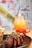 A plate of glazed ribs garnished with chopped green onions and white flowers is paired with a side of fries in a metal basket. In the background, a vibrant gradient drink in a mason jar with a straw features layers of orange, yellow, and red, styled with blueberries and a garnish.