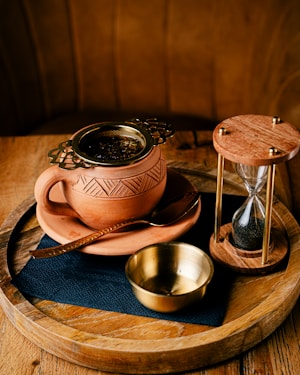 A beautifully handcrafted terracotta cup with intricate patterns holds brewing tea leaves in a metal strainer. Accompanying it is a small brass cup and a rustic hourglass, all placed on a wooden tray, creating a cozy and artisanal setting.
