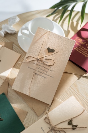 A beautifully arranged scene featuring greeting cards tied with twine, placed on a wooden table. The central card is cream-colored with an embossed message and a small decorative heart. Nearby, there is a white teacup and saucer, accompanied by a couple of cards in green and maroon shades, and some leafy branches adding a natural touch.