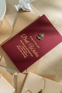 A maroon greeting card with gold text lies on a wooden surface. The text expresses warm wishes for joy and happiness. The card is adorned with a small metallic rose decoration. Surrounding the card are brown and cream-colored envelopes, one of which is partly visible. The scene is softly illuminated, casting gentle shadows.