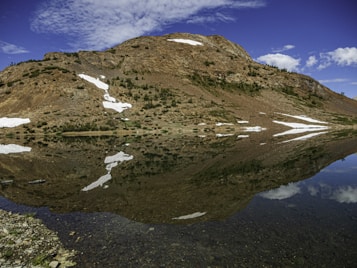 A rocky mountain landscape with patches of snow reflects perfectly in a calm, clear lake. The sky is a vibrant blue with scattered clouds, and the natural scene conveys a sense of tranquility and pristine beauty.