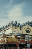An urban street scene featuring a multi-story building with signs written in Vietnamese. The structure has a weathered facade, adorned with vibrant red vines. Large lettering spelling 'Hanoi' is prominently displayed on the roof. Traditional architecture elements are visible, including arched windows. Street lamps appear in the foreground, along with traffic signage.
