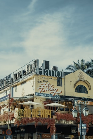 An urban street scene featuring a multi-story building with signs written in Vietnamese. The structure has a weathered facade, adorned with vibrant red vines. Large lettering spelling 'Hanoi' is prominently displayed on the roof. Traditional architecture elements are visible, including arched windows. Street lamps appear in the foreground, along with traffic signage.