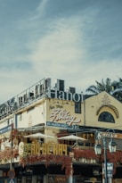 An urban street scene featuring a multi-story building with signs written in Vietnamese. The structure has a weathered facade, adorned with vibrant red vines. Large lettering spelling 'Hanoi' is prominently displayed on the roof. Traditional architecture elements are visible, including arched windows. Street lamps appear in the foreground, along with traffic signage.
