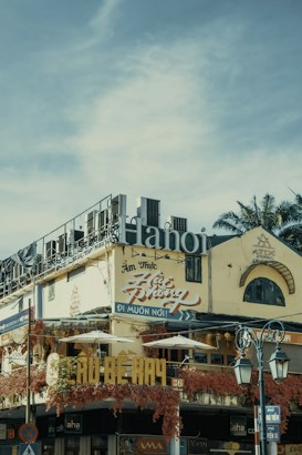 An urban street scene featuring a multi-story building with signs written in Vietnamese. The structure has a weathered facade, adorned with vibrant red vines. Large lettering spelling 'Hanoi' is prominently displayed on the roof. Traditional architecture elements are visible, including arched windows. Street lamps appear in the foreground, along with traffic signage.