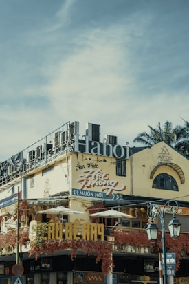 An urban street scene featuring a multi-story building with signs written in Vietnamese. The structure has a weathered facade, adorned with vibrant red vines. Large lettering spelling 'Hanoi' is prominently displayed on the roof. Traditional architecture elements are visible, including arched windows. Street lamps appear in the foreground, along with traffic signage.