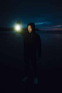 A person stands in a dimly lit area holding a glowing lantern in one hand. The background reveals a serene, dark landscape, suggesting either dusk or dawn, with the faint outline of water and sky.