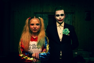 Two individuals are dressed in costumes resembling popular fictional characters. The person on the left has long blonde hair styled in pigtails, wearing a colorful jacket and a T-shirt. The person on the right wears a dark suit with a white shirt, accented with face paint resembling a clown or jester.