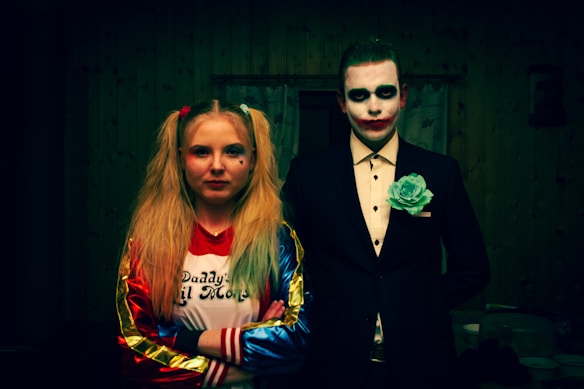 Two individuals are dressed in costumes resembling popular fictional characters. The person on the left has long blonde hair styled in pigtails, wearing a colorful jacket and a T-shirt. The person on the right wears a dark suit with a white shirt, accented with face paint resembling a clown or jester.