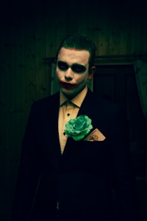 A person appears with face paint resembling a clown, characterized by dark eye makeup and red lips. They wear a formal black suit with a pale shirt, accessorized with a large green flower and a visible playing card in the breast pocket. The background is a dimly lit wooden wall.