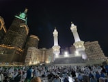 A nighttime scene featuring majestic architecture illuminated by lights. Tall towers with intricate designs stand prominently, while a large crowd of people, many dressed in white clothing, gather in the open space below. The ambiance suggests a significant cultural or religious gathering.