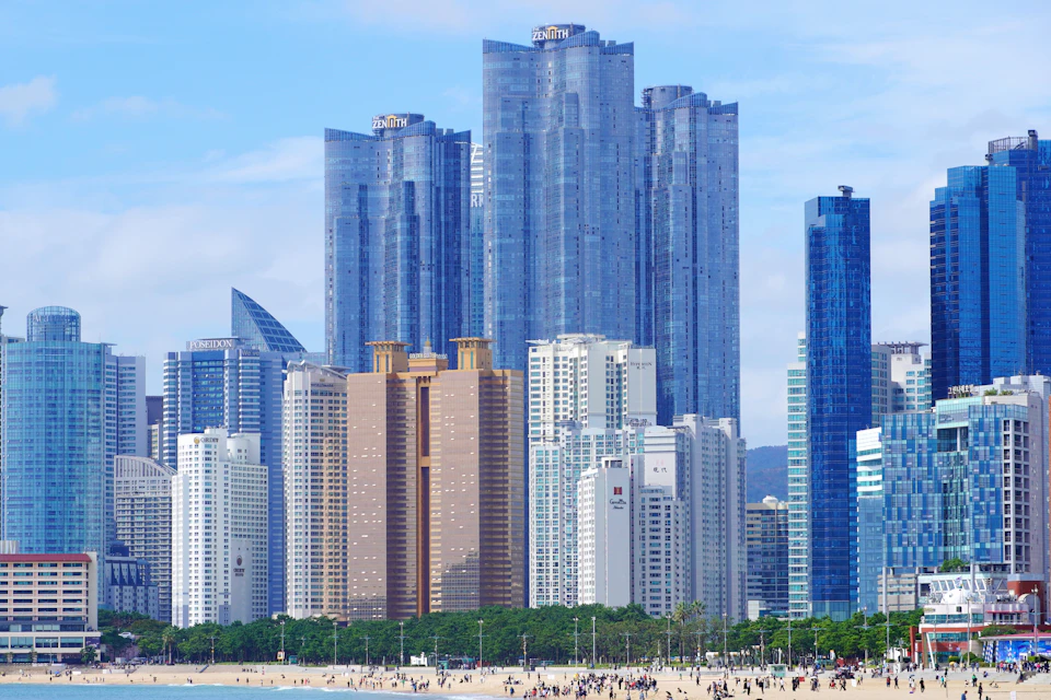 The image depicts a bustling cityscape with numerous tall buildings, predominantly white and gray in color, reaching towards the sky. The tallest building is located on the left side of the frame, while another skyscraper can be seen on the right side. In the foreground, there are several smaller buildings that add depth to the scene. The beach is visible in the background, with people walking along its edge and enjoying a day at the shoreline.