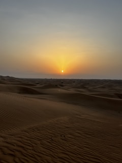 A serene desert landscape featuring vast, undulating sand dunes under a clear sky. The sun is setting on the horizon, casting a warm golden glow across the sand and sky, creating a peaceful and tranquil atmosphere.